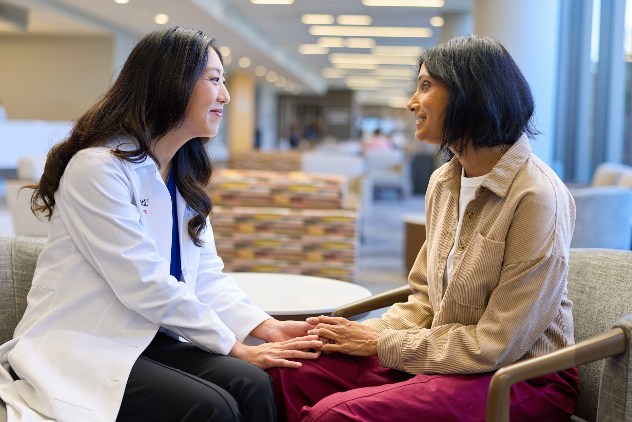 Lindsay Kuroki, MD, MSCI, wearing a WashU Medicine lab coat, smiles as she holds hands with a seated patient in a lobby area.