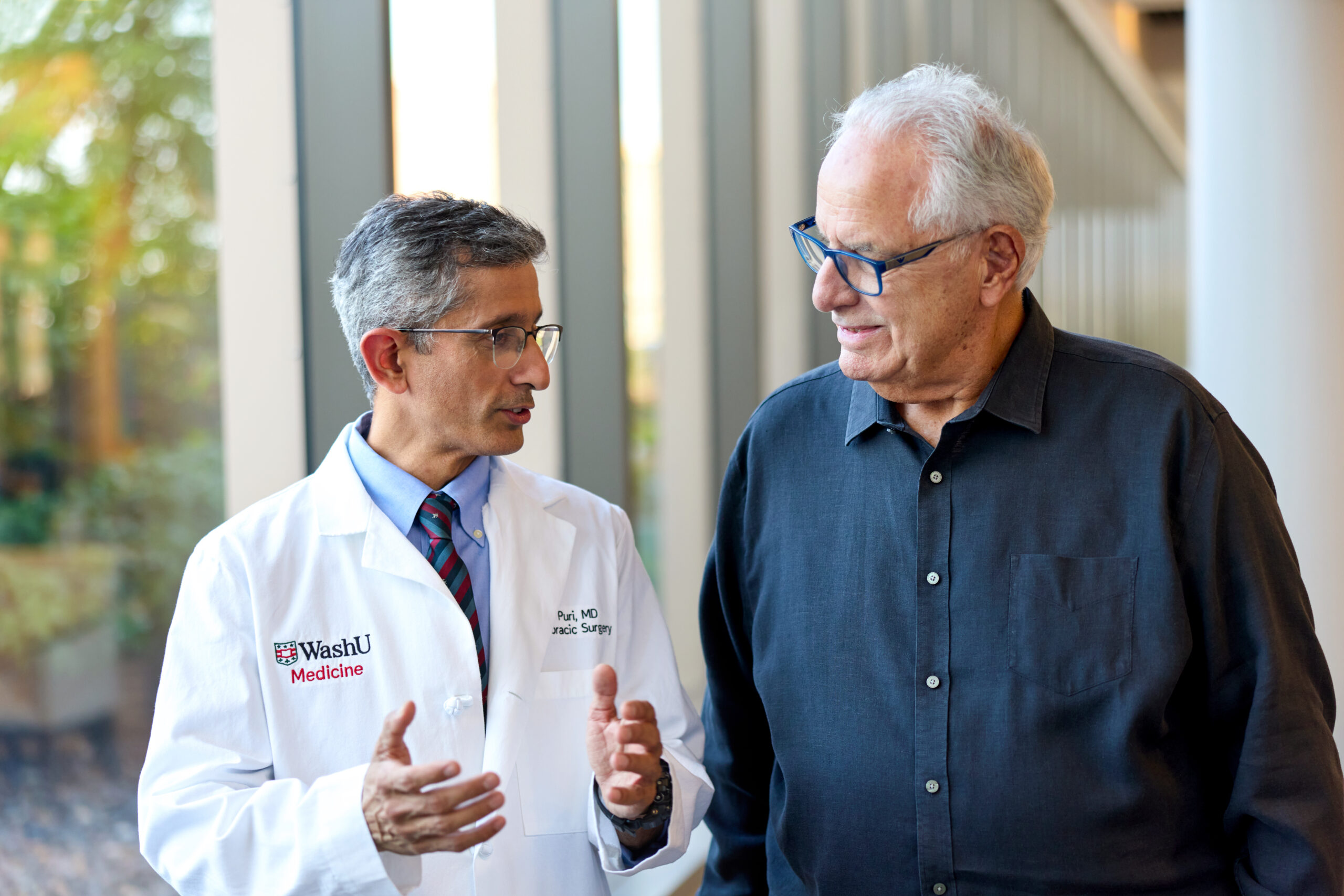 Dr. Varun Puri, MD, MSCI, wearing a WashU Medicine lab coat, gestures with his hands while speaking to a patient wearing a dark blue button-down shirt.