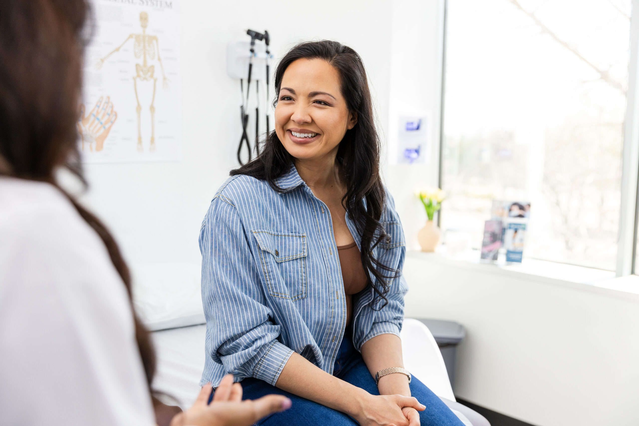 The mid adult female patient sits on the examination table and smiles while listening to encouraging news from the doctor.