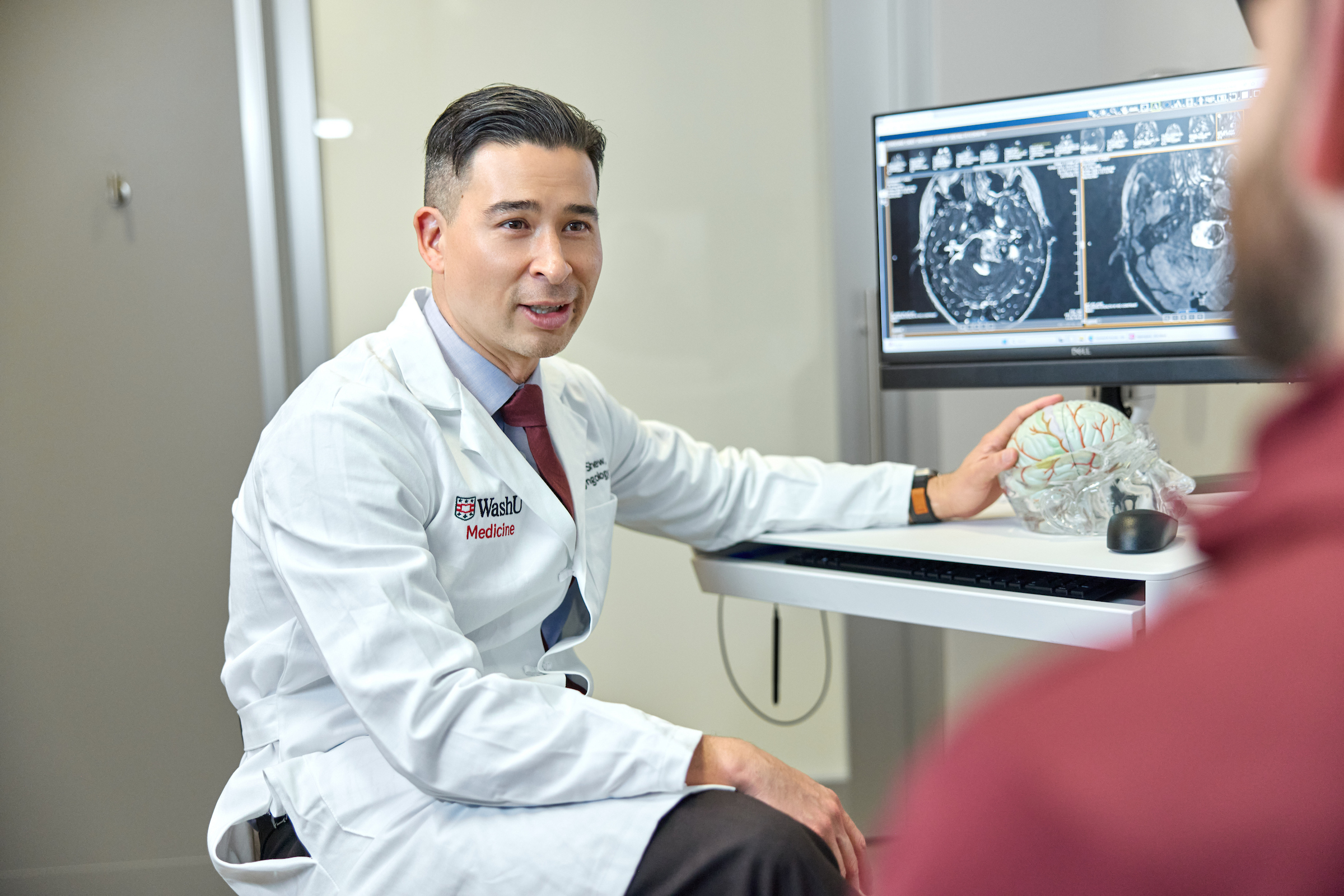 A doctor holds a replica of a brain with image mapping on the computer behind him while speaking with a patient