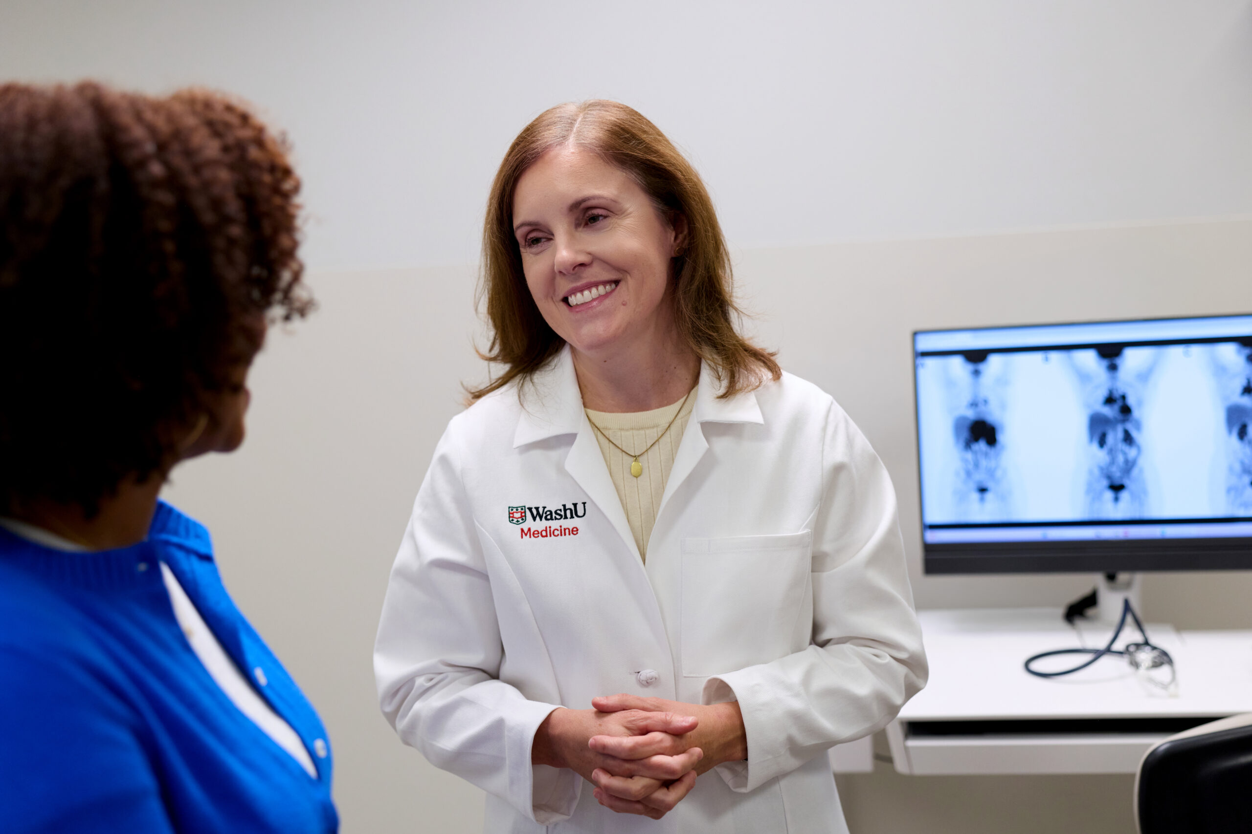 Dr. Amanda Cashen, MD, wearing a WashU Medicine lab coat, smiles while speaking with a patient in an exam room with medical scans on a screen in the background.