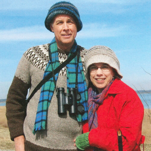portrait of a man and his wife outside with a lake behind them and binoculars draped over the man's shirt