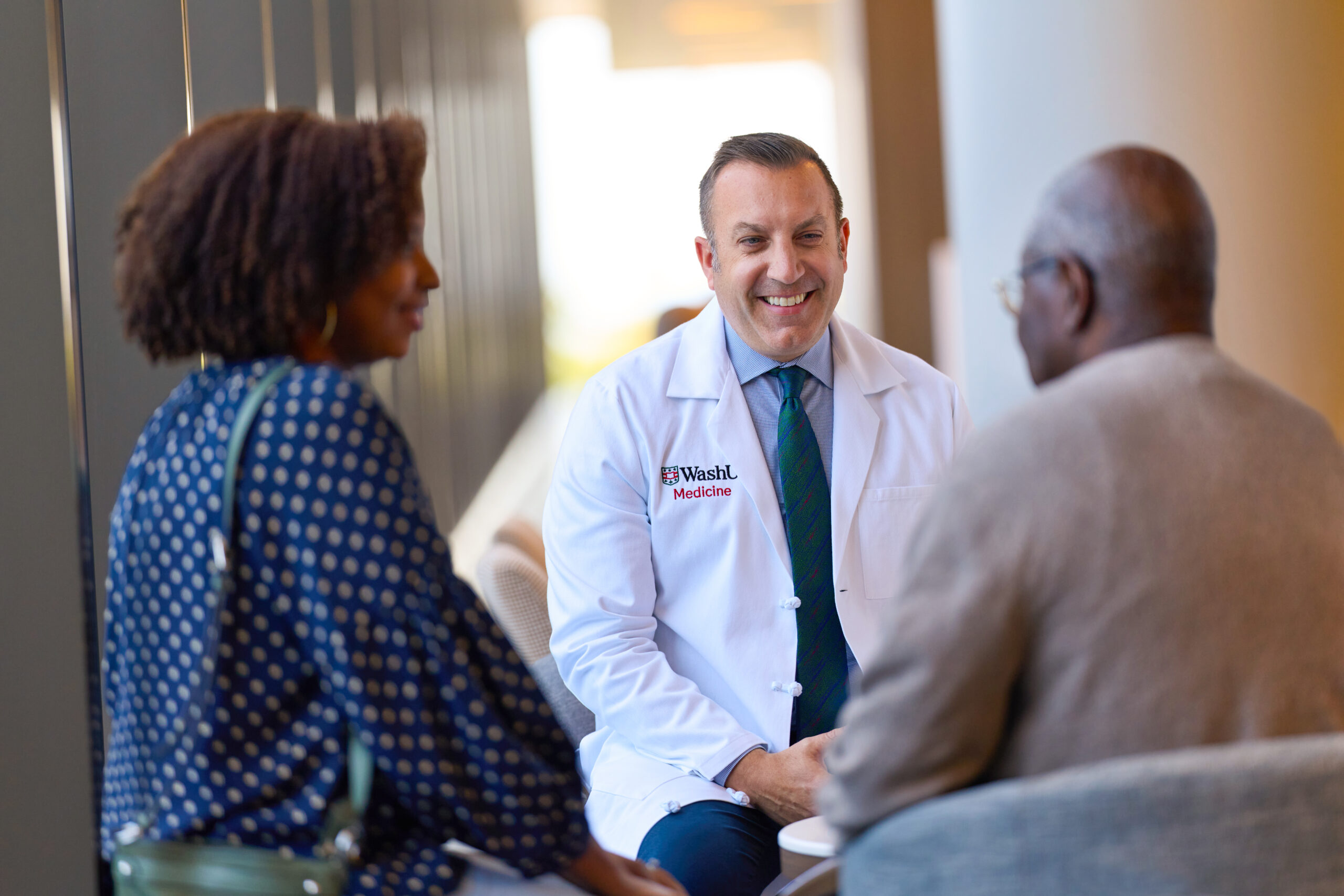 Gregory Vlacich, MD, PhD wearing a WashU Medicine lab coat, smiles while conversing with a patient and a companion seated across from him.