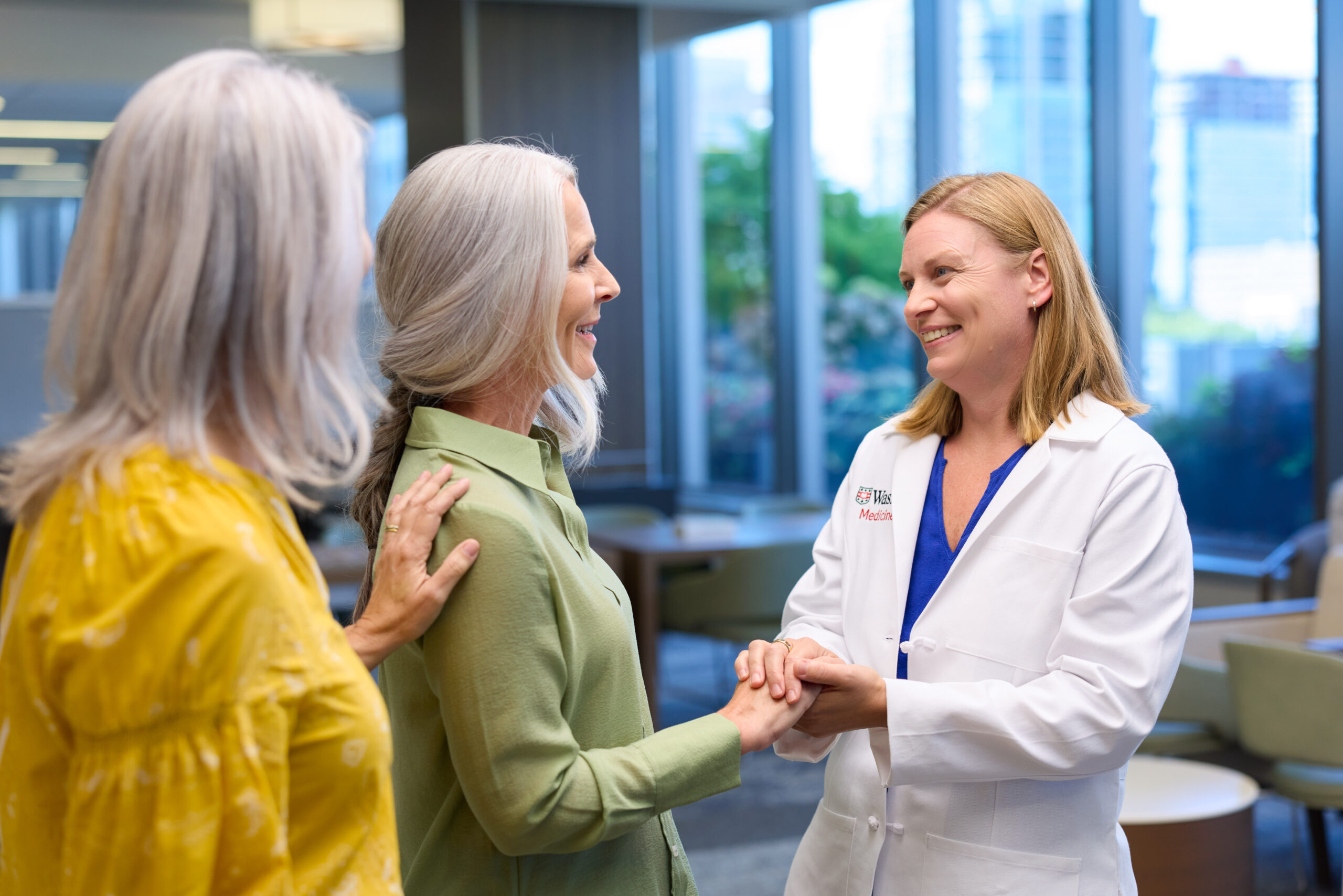 Dr. Lindsay Peterson, MD, MSCR, wearing a WashU Medicine lab coat, smiles while holding hands with a patient standing beside another woman near large windows.