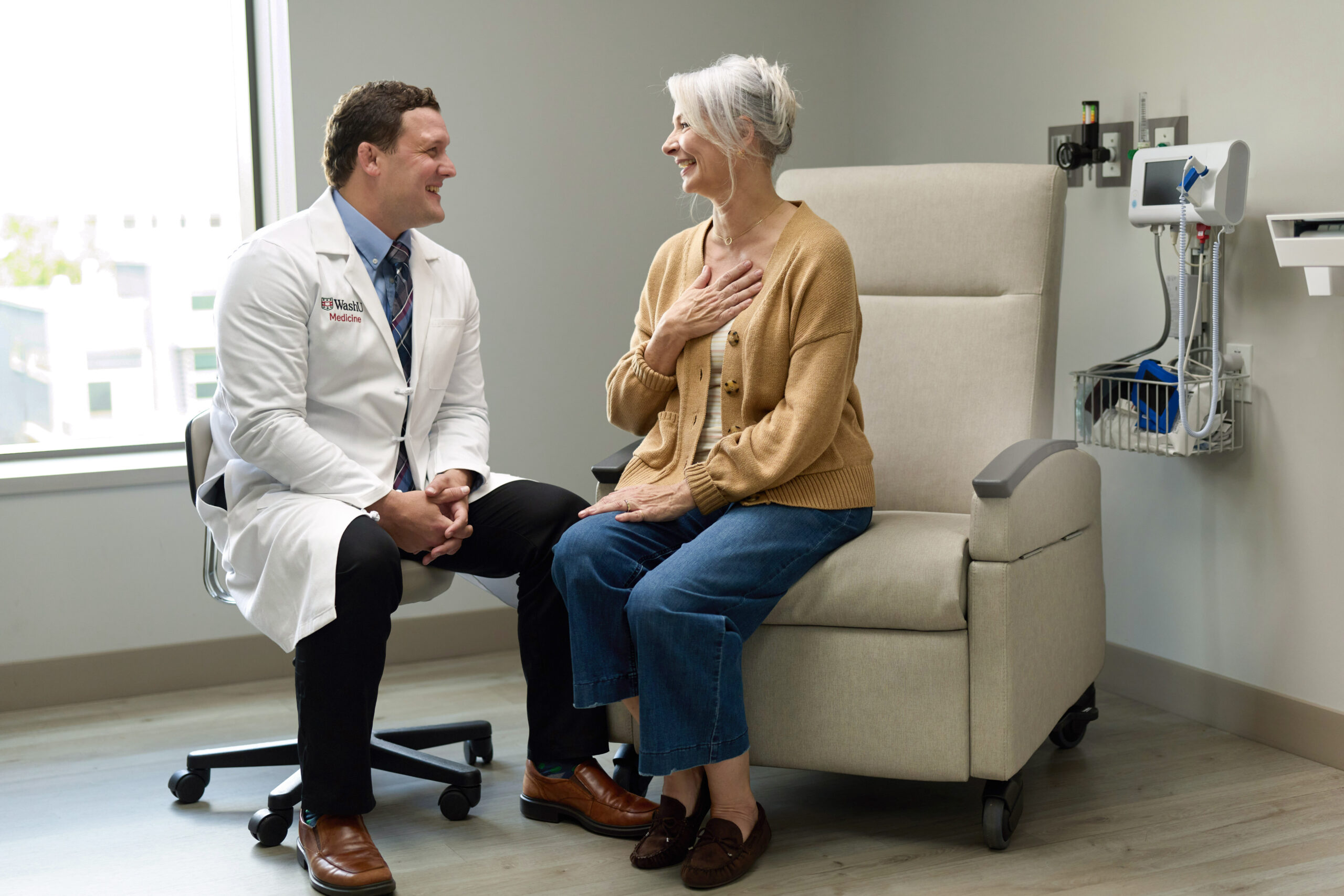 Dr. Michael Waters, MD, PhD, wearing a WashU Medicine lab coat, smiles as he speaks with a patient seated in a medical recliner.