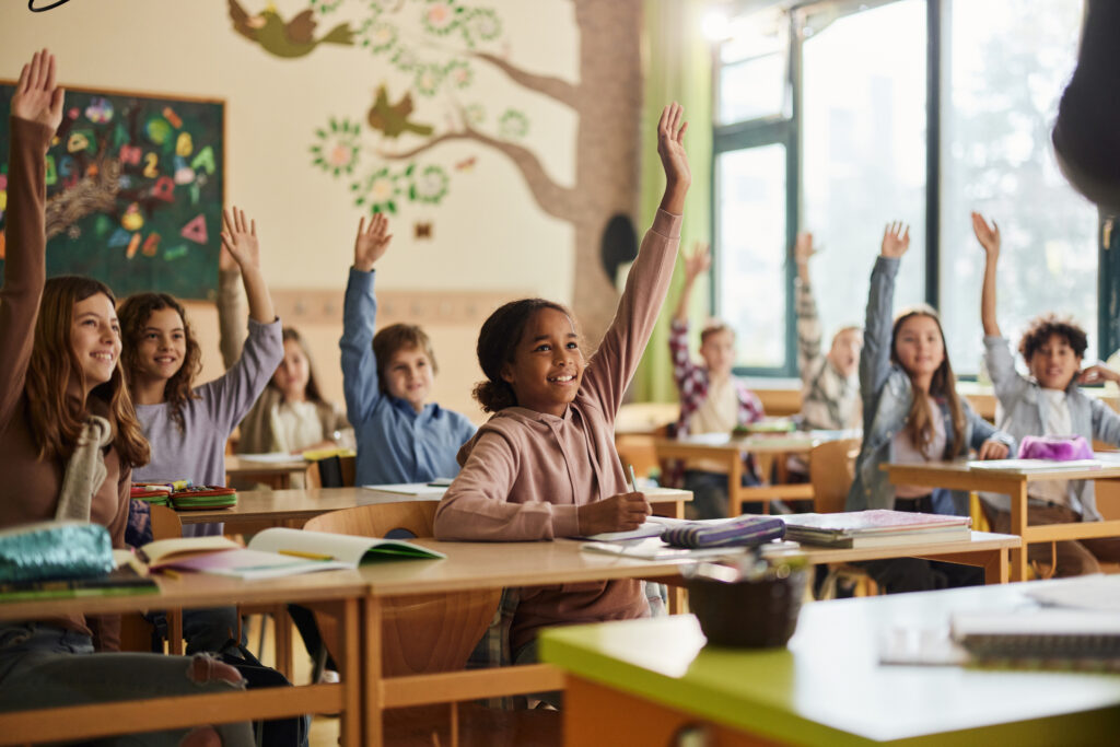 Elementary schoo students are shown raising their hands in a classroom