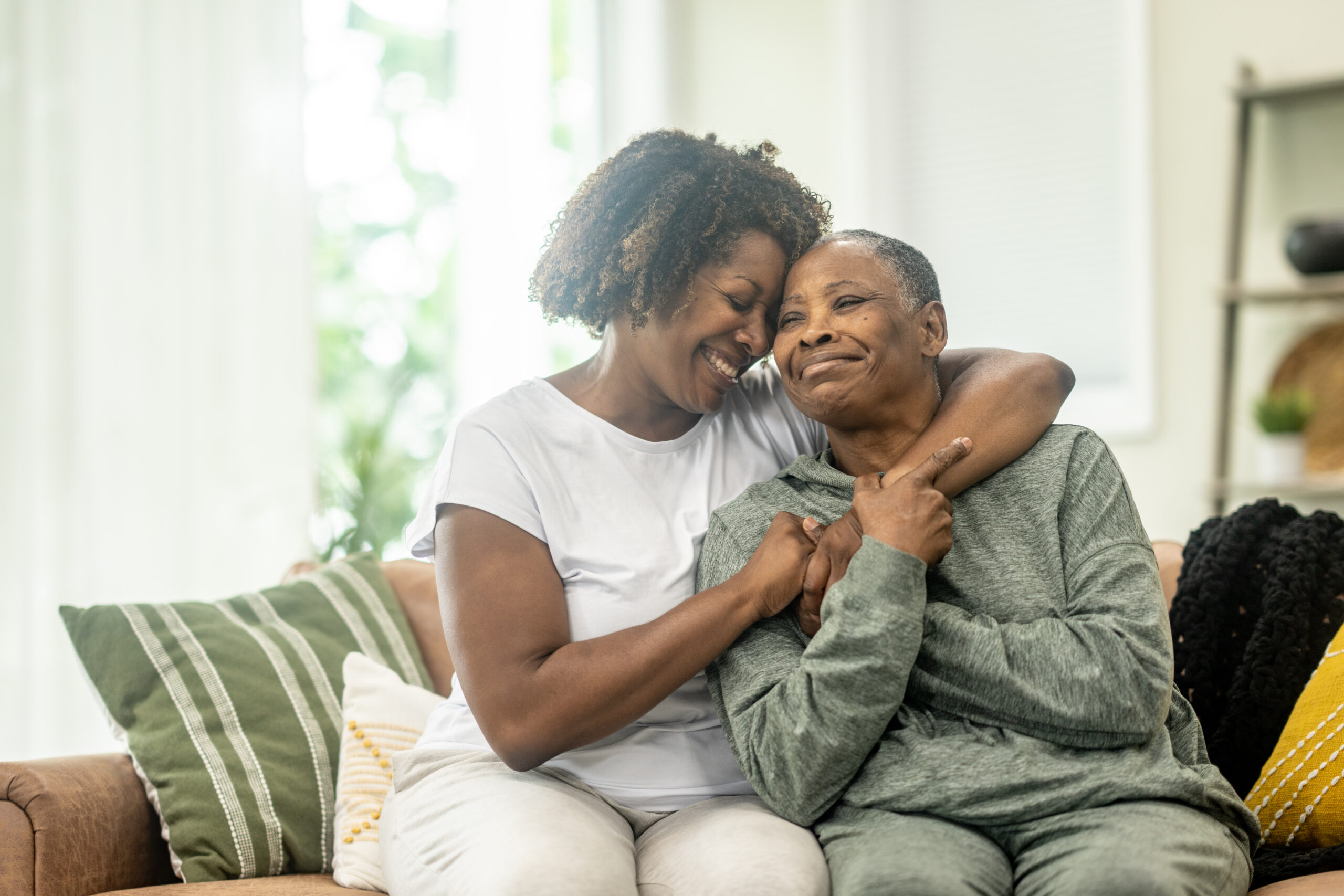 Two women sit side-by-side on a couch, with one woman leaning in to hug the other from behind as both smile.