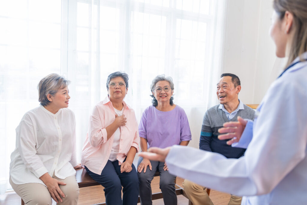 A healthcare professional in a lab coat gestures while speaking to a group of four older adults seated along a bench in a bright room.