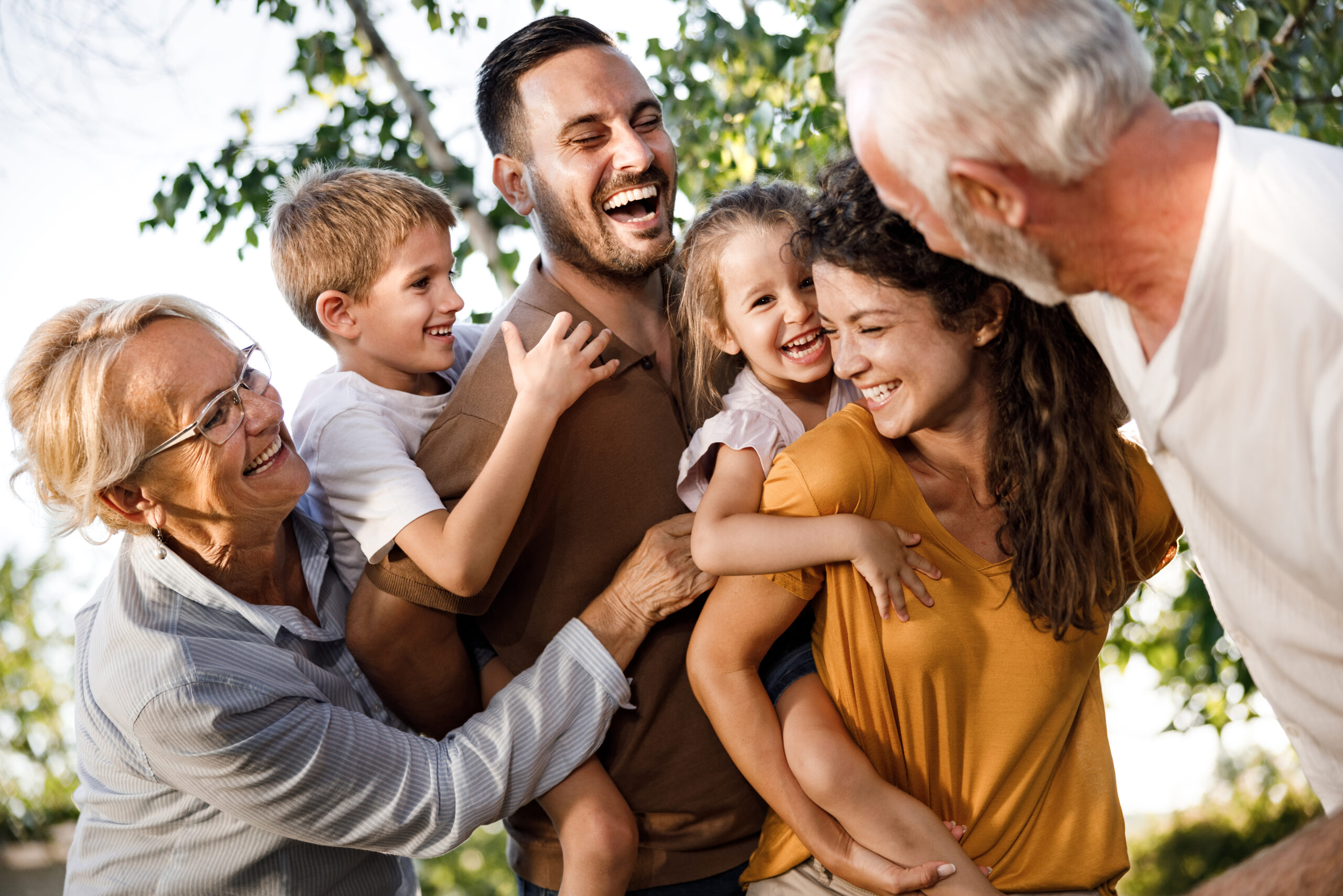 A multi-generational family smiles and laughs together outdoors while two adults give piggyback rides to young children.