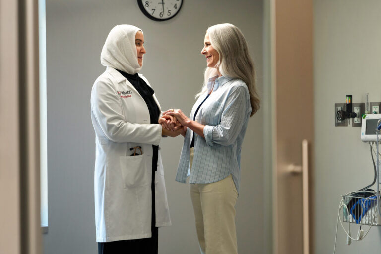 Nusayba Bagegni, MD wearing a WashU Medicine lab coat, holds hands with a patient while smiling in a clinical room.