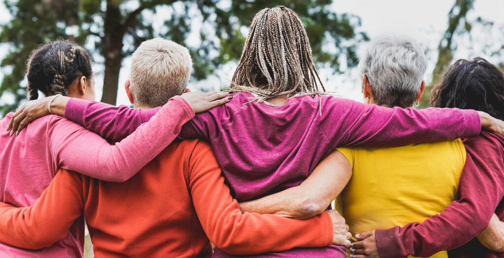Five people seen from behind stand side-by-side with their arms wrapped around each other's shoulders outdoors.