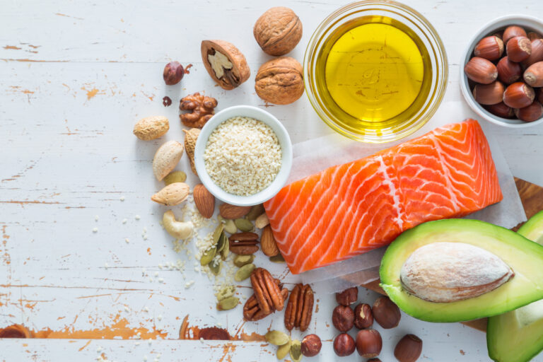 An overhead display of healthy food items, including a salmon fillet, avocado half, oil, seeds, and various nuts on a white wooden table.