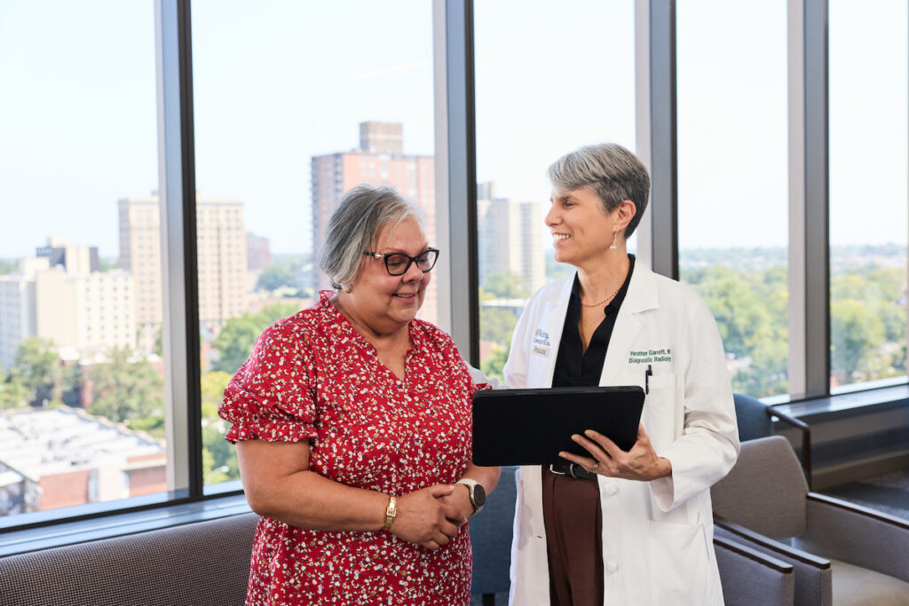 A healthcare professional in a white coat holds a tablet and speaks with a patient in a red floral dress, both smiling, in a bright room with large windows overlooking a cityscape.