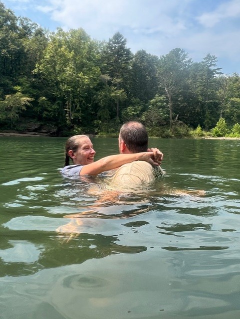 Rick and Dianna Sparrow smiling and embracing in a natural body of water surrounded by trees.   