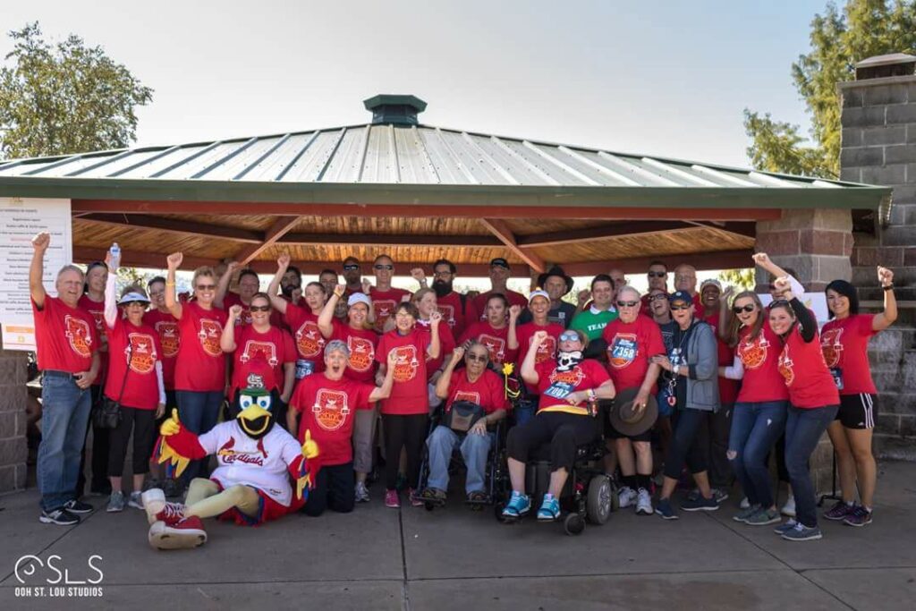 A large group of people in red shirts poses outdoors under a pavilion alongside a bird mascot.